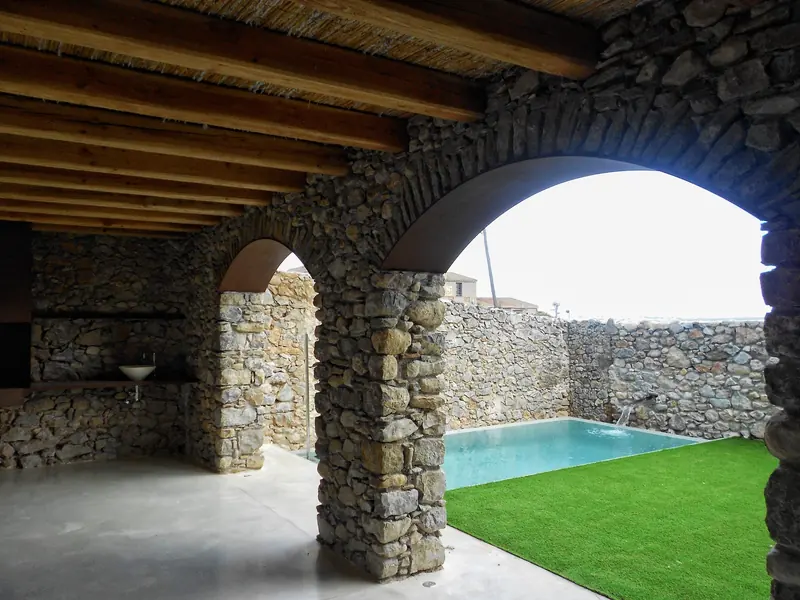 Porxo de pedra amb arcs i bigues de fusta obert a una piscina i gespa